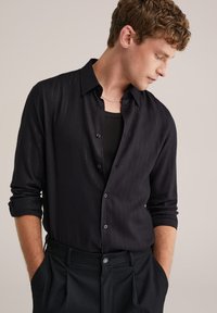 Must värviline särk, millel on õrnad vertikaalsed triibud, pikad varrukad rullitud mansettides, koos musta lõhikuga ja kohandatud pükstega.