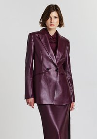 Donna che indossa un blazer doppiopetto in pelle viola scuro sopra una camicia e una gonna coordinate, in piedi contro uno sfondo chiaro e semplice.