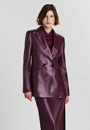Femme portant un blazer en cuir double boutonnage violet foncé sur une chemise et une jupe assorties, se tenant devant un fond clair.