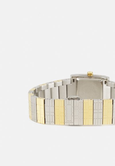 Hõbedane ja kuldne metallkell, millel on ristkülikuline korpus, tekstureeritud linkide ja vahelduvate halli ja kollase sektsioonidega, ning kuldne kroon.