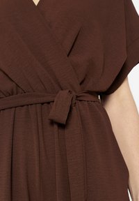 Bruine wikkeljurk van lichtgewicht stof, met korte mouwen, een strikdetail in de taille en plooien voor extra textuur.