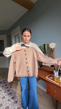 Frau hält eine beige, knöpfbare Strickjacke an einem Kleiderbügel in einem Raum mit einer Holzkommode, einem Spiegel und einer dekorativen Blume.
