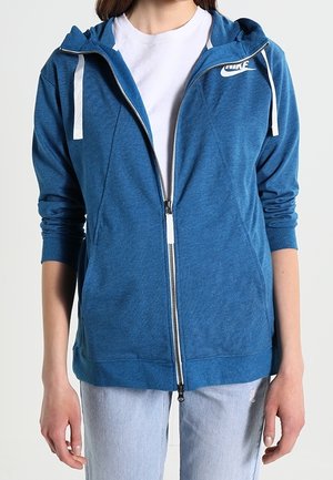 Persoon draagt een blauwe Nike-hoodie met rits over een wit shirt, gecombineerd met lichtblauwe spijkerbroek, staand tegen een effen achtergrond.