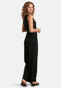 Jumpsuit nero a coste con design senza maniche, vita cintrata e pantaloni a gamba larga. Indossato con sandali neri con tacco.