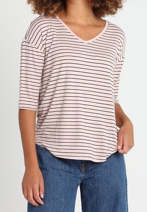 Kobieta ubrana w jasnoróżową bluzkę z dekoltem w serek i cienkimi czarnymi poziomymi paskami oraz niebieskie dżinsy, stojąca z jedną ręką w kieszeni.