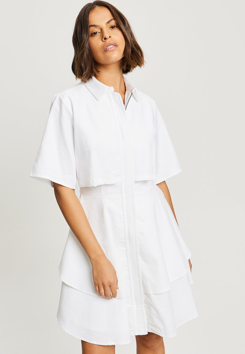 Robe blanche avec un col, des manches courtes et un ourlet en couches. Fabriquée en tissu lisse, elle possède une fermeture éclair à l'avant et une coupe ajustée.