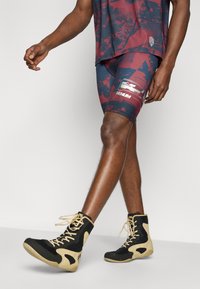 Αθλητικές βερμούδες σε μπορντό με διακριτικό σχέδιο, σε συνδυασμό με μαύρα και καφέ μπότες προπόνησης που διαθέτουν υλικά από δίχτυ και καουτσούκ.