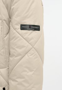 Beige geborduurde jas met een zachte textuur, voorzien van een zwart stoffen label met "FRIEDA FREDDIES NEW YORK" op de bovenarm.