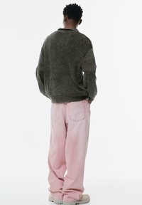 Persoon in een donkergrijze oversized trui, lichtroze wijde jeans en lichtbeige laarzen, staande met de handen in de zakken, met de rug naar voren gericht.