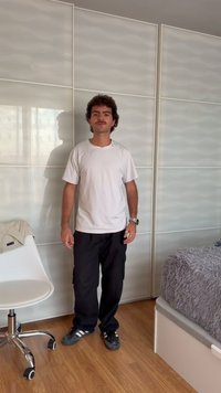 Homme avec moustache portant un t-shirt blanc, un pantalon noir et des baskets, debout dans une chambre avec une chaise blanche et un lit avec une couverture grise.