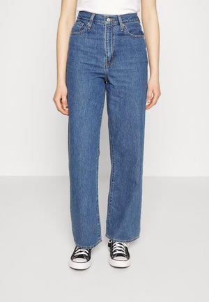 Persoon draagt een hoge taille blauwe rechte jeans en zwarte lage sneakers met witte veters, staand tegen een lichte achtergrond.