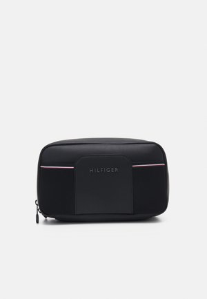 Rektangulær sort toilettaske med frontlomme, lynlåslukning og diskrete røde og hvide striber over "Hilfiger"-mærket.