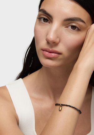 Młoda kobieta o ciemnych włosach, ubrana w biały, bezrękawnik, z czarną bransoletką z koralików z małym złoto-czarnym zawieszką na nadgarstku.