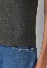 Grijze gebreide trui met ribbelzoom en klein metalen logo detail aan de onderkant, gecombineerd met blauwe denim jeans met een zichtbare zak.