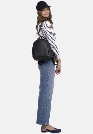 Schwarzer Leder-Rucksack mit runder Form, verstellbaren Riemen und minimalem Beschlag. Das Modell trägt ein gestreiftes Langarmshirt und blaue Jeans.