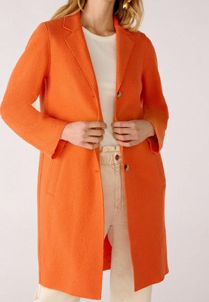 Kvinde iført en knaldorange, midt-lang frakke med hakkede slag over en hvid ribbet top og lyse beige bukser, der holder i frakkens forkant.