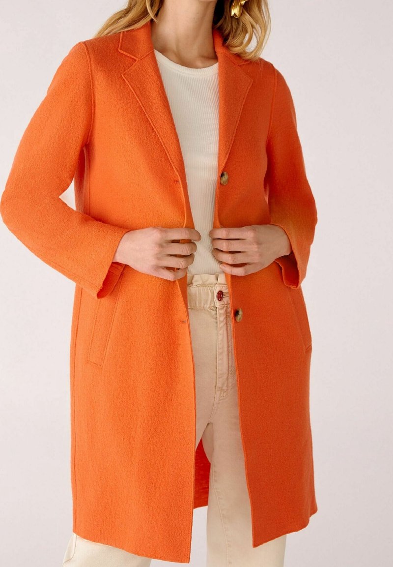 Femme portant un manteau mi-long orange vif à revers crantés sur un haut blanc côtelé et un pantalon beige clair, tenant les bords avant du manteau.