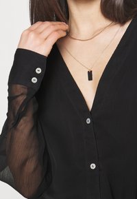 Žena v černé blůze s průhlednými rukávy a dvěma zlatými náhrdelníky, jedním řetízkem a jedním s obdélníkovým černým přívěskem, upravující řetízkový náhrdelník.