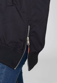 Zwarte bomberjack met een zilveren metalen rits aan de zijkant, geribde manchetten en een gladde textuur. De voering heeft een oranje accent.