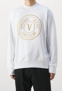 Vit sweatshirt med ett guldbroderat logodesign på framsidan. Mjuk tyg, ribbad manschetter och nederkant, avslappnad passform, minimalistisk stil.