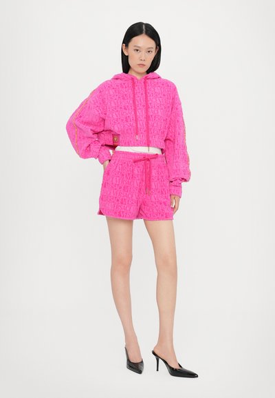 Svetlo roza skrajšana kapuca in kratke hlače z embosiranim logotipom, elastičnim pasom in zlatimi dodatki, v kombinaciji s črnimi petami.