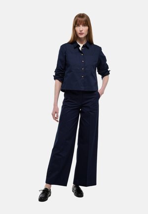 Femme debout, une main dans la poche, portant une chemise bleu marine boutonnée avec les manches retroussées, un pantalon large assorti et des chaussures noires à lacets.