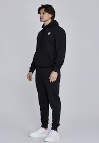 Czarna bluza z kapturem i dopasowane dresy z przednią kieszenią, z małym białym logo, w zestawie z białymi trampkami. Gładka tekstura materiału.