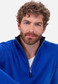 Pull à col zippé bleu en tissu tricoté doux, avec des accents noirs et une finition texturée, associé à une couche de base blanche.