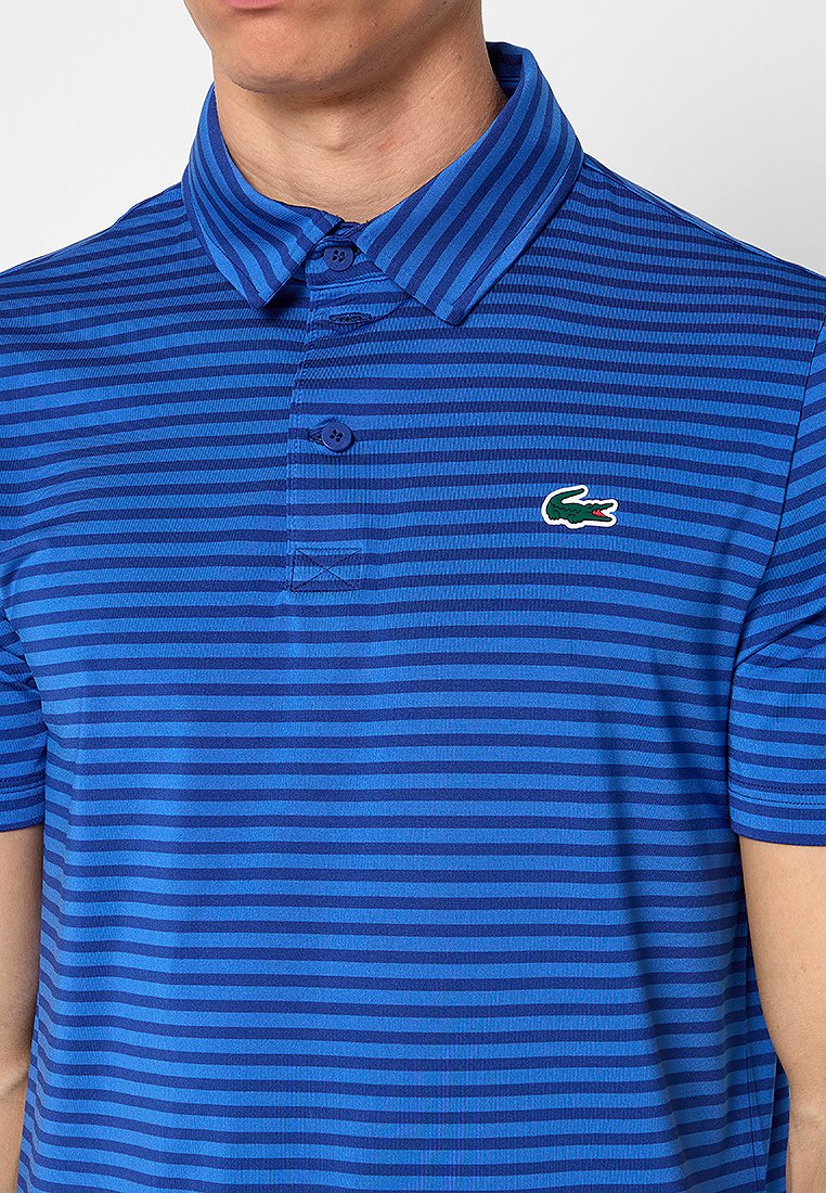Chemise polo bleue avec des rayures horizontales de différentes teintes ; elle possède un col à côtes, une patte de boutonnage à trois boutons et un logo de crocodile vert.