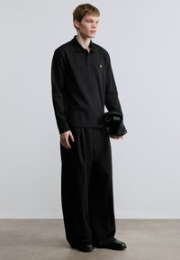 Μαύρο πόλο πουκάμισο μακρυμάνικο με μικρό λογότυπο, χαλαρή εφαρμογή και βαμβακερό ύφασμα. Φορώντας το με φαρδιά μαύρη παντελόνια και κρατώντας μαύρη τσάντα.