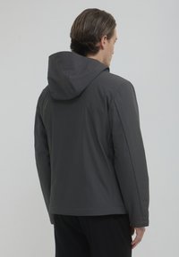 Ein Mann, der mit dem Rücken zur Kamera steht, trägt eine dunkelgraue Kapuzenjacke und schwarze Hosen vor einem schlichten hellen Hintergrund.