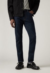 Homme portant un jean slim bleu foncé, des chaussures en cuir noires, une chemise grise et une veste noire, debout avec une main dans la poche.