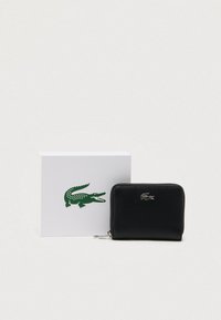Cartera con cremallera alrededor, de cuero negro texturizado, con logo de cocodrilo plateado junto a una caja blanca con logo de cocodrilo verde sobre un fondo liso.