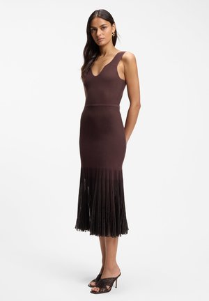 Kobieta stojąca, ubrana w bezrękawową, dopasowaną ciemnobrązową sukienkę z plisowanym dołem i ciemne, otwarte sandały na obcasie na białym tle.