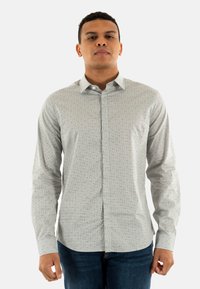 Chemise boutonnée de couleur claire avec un petit motif géométrique, manches longues et col classique. Portée avec un jean en denim bleu.