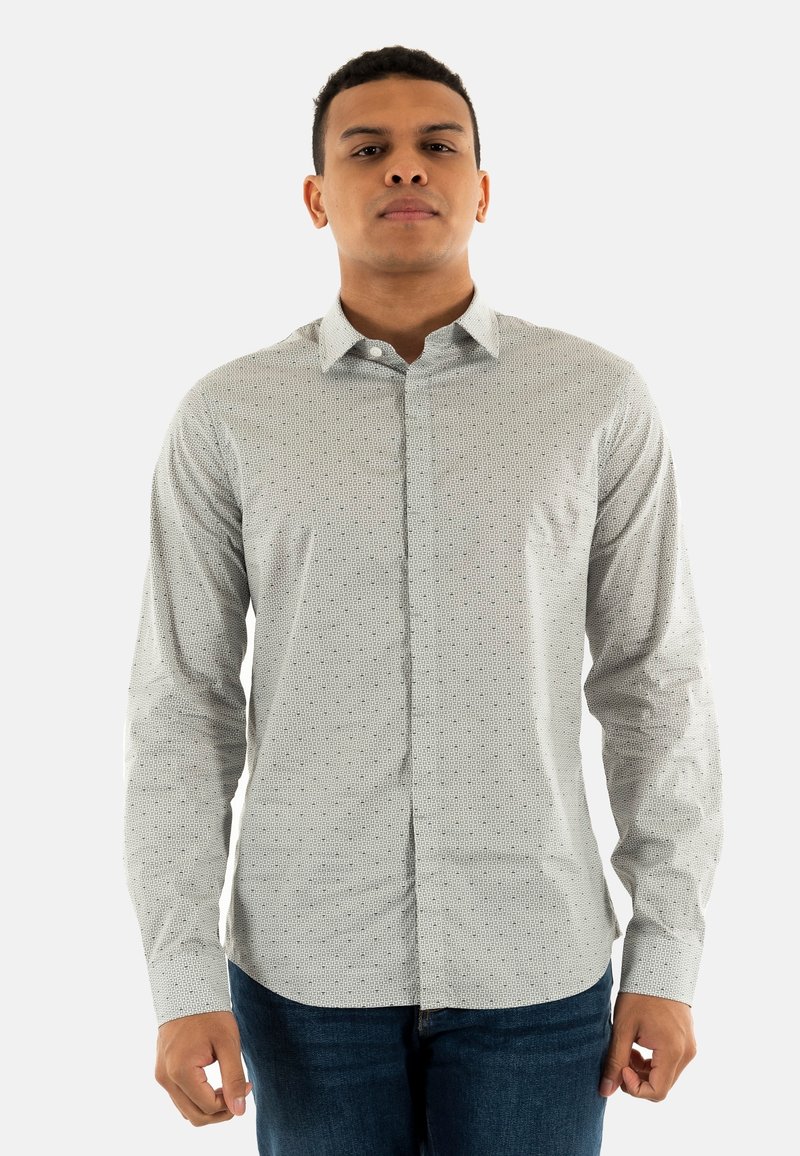 Chemise boutonnée de couleur claire avec un petit motif géométrique, manches longues et col classique. Portée avec un jean en denim bleu.