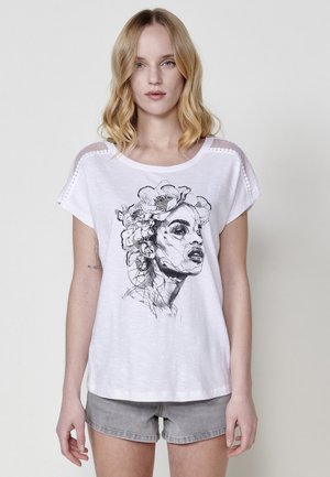 T-shirt en coton blanc à manches courtes, présentant un croquis graphique noir d'un visage de femme orné de fleurs. Accents texturés aux épaules.