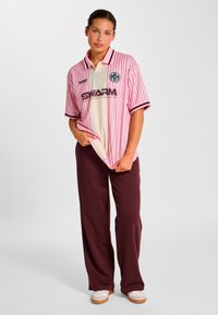 Pinkstribet poloshirt med sorte accenter og et hvidt centerpanel, som har logoet "SWARM International". Kombineret med løse burgunderrøde bukser.
