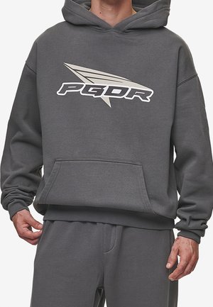 Personne portant un sweat à capuche gris foncé et un pantalon assorti, le sweat à capuche arbore un logo stylisé "PGDR" en blanc et beige sur la poitrine.