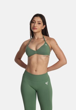 Ung kvinde med langt mørkt hår iført grøn sports-bh og leggings, stående med hænderne bag ryggen på hvid baggrund.