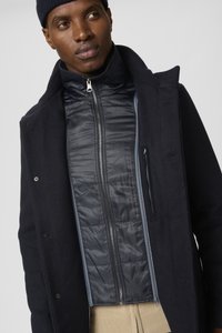 Giubbotto imbottito blu navy con un interno liscio e lucido, indossato sotto un cappotto di lana scuro con colletto e tasche con zip.