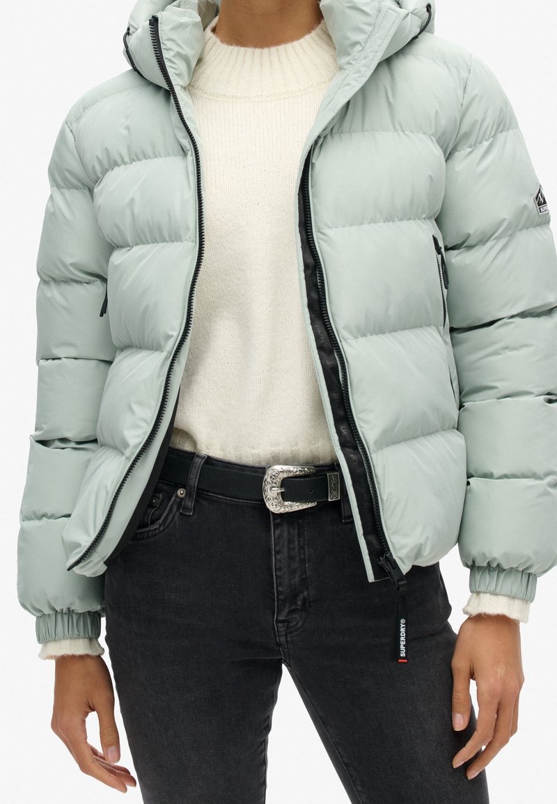 Personne portant une doudoune gris clair par-dessus un pull blanc, associée à un jean noir et une ceinture noire avec une boucle argentée.