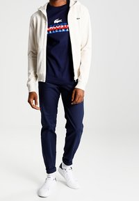 Sudadera con cremallera beige sobre una camiseta navy que presenta un gráfico en rojo y blanco, combinada con pantalones navy y zapatillas blancas.