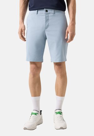 Mężczyzna w jasnoniebieskich szortach, białych tenisówkach z zielonymi sznurowadłami i białych skarpetach do kostek, stojący na jednolitym tle.