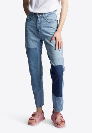 Person iført høj talje patchwork lys og mørkeblå jeans med pink åbne tå-sandaler, stående mod en hvid baggrund.