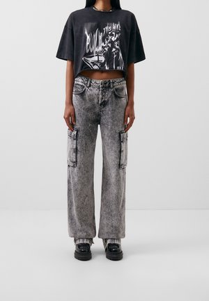 Kvinde iført sort kort grafisk t-shirt, høj talje grå acid wash cargo jeans med sidelommer og sorte plateau sko.