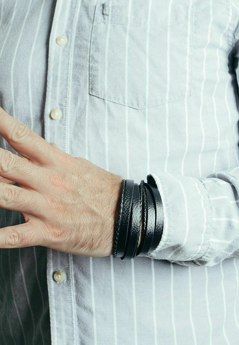 Bracciale in pelle nera con multiple fasce sottili sovrapposte. Indossato su una mano vicino a una camicia a righe grigio chiaro.