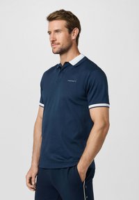 Homme portant un polo bleu marine avec un col blanc et des rayures sur les manches, debout, une main dans la poche, faisant face à gauche.