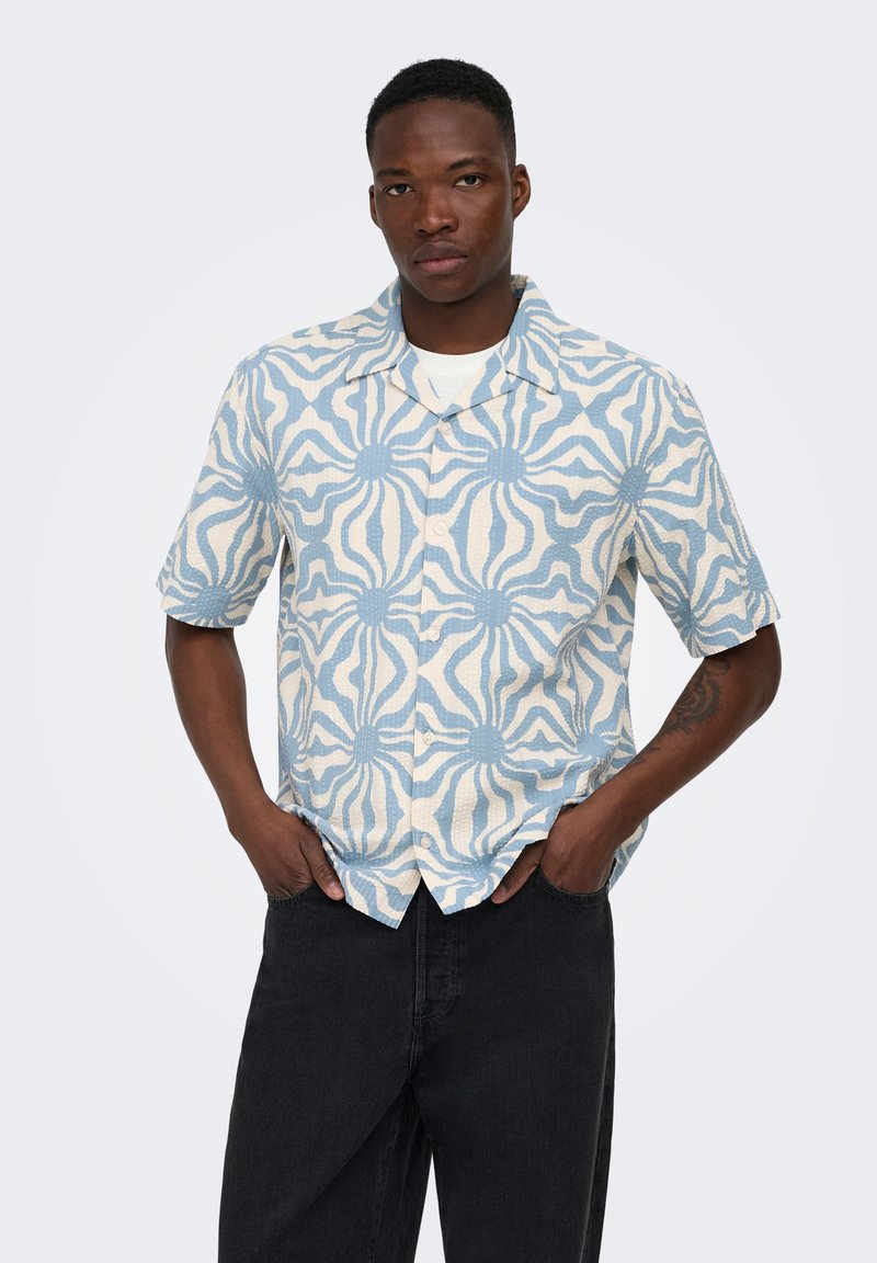Homme portant une chemise à manches courtes boutonnée avec un motif bleu et blanc, les mains dans les poches d’un pantalon noir, debout devant un fond uni.
