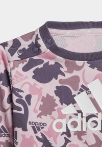 Sweatshirt rose et violet avec un motif de dinosaure, doté d'un col côtelé et de boutons pression. Logo Adidas blanc sur le devant.